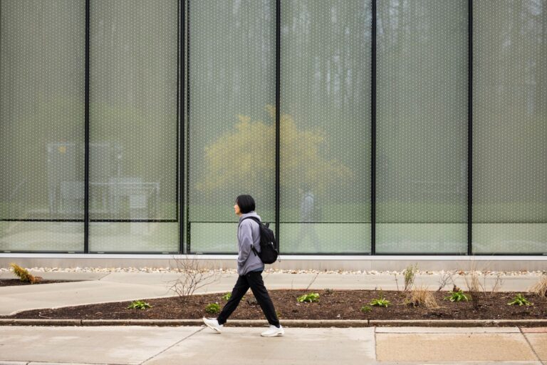An Asian woman walks past a building with tall, rectangular floor-to-ceiling windows with black frames. She is dressed in black pants, a grey hoodie and has a black backpack.