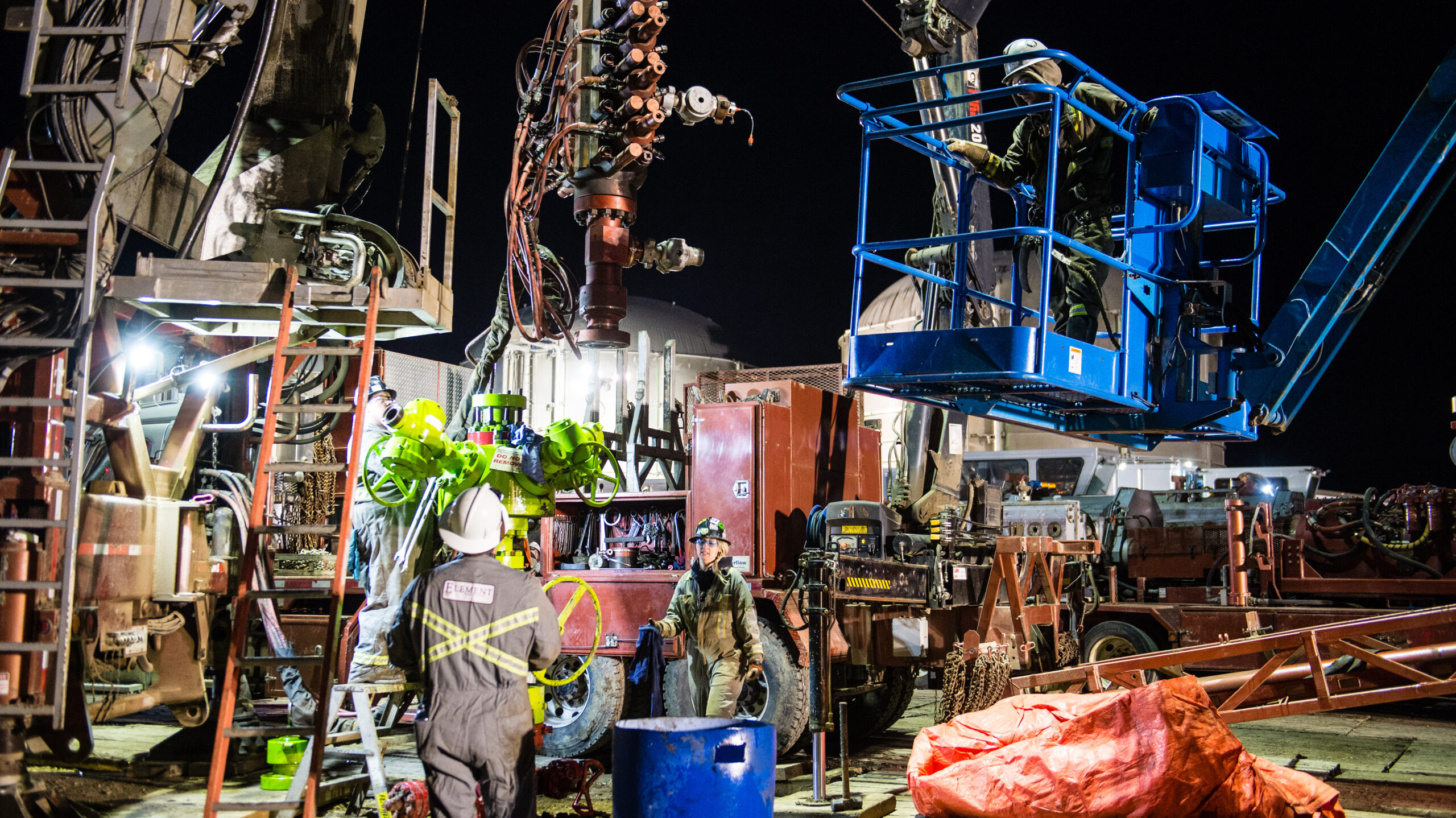 Three workers in grey coveralls and hardhats work outside at night on a site filled with machinery, ladders, industrial vehicles, metal lifts, and tarps covering things.