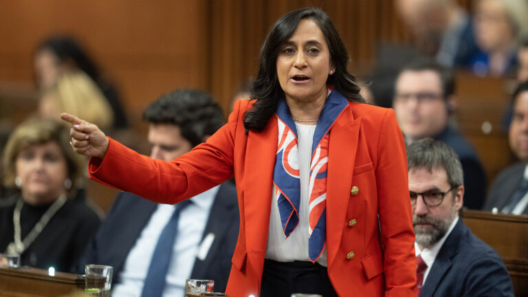Anand is standing with one arm out, pointing to something. She is wearing a red blazer, white shirt and a scarf that is blue, red and white.