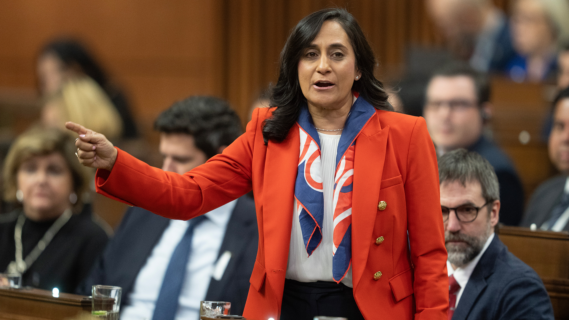 Anand is standing with one arm out, pointing to something. She is wearing a red blazer, white shirt and a scarf that is blue, red and white.
