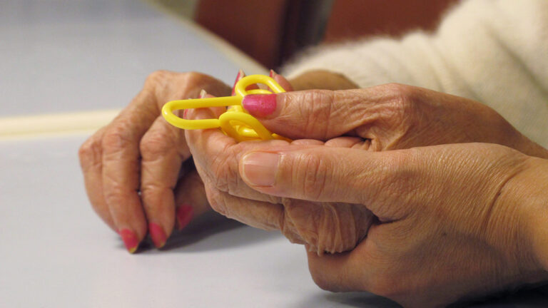 Two sets of hands, one set with nails painted pink, hold a small yellow chain-link puzzle against a grey table.