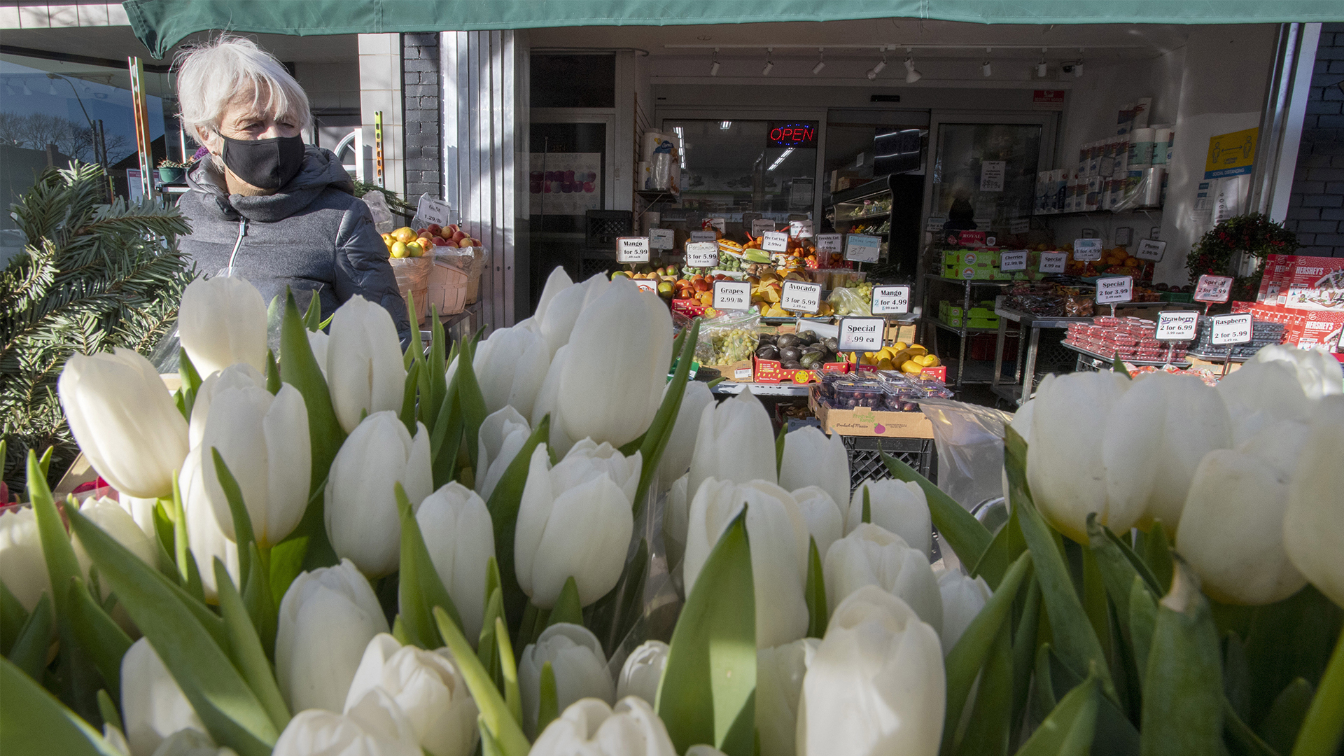 A woman in a mask shops in front of a large display of white tulips with a small grocery store in the background.