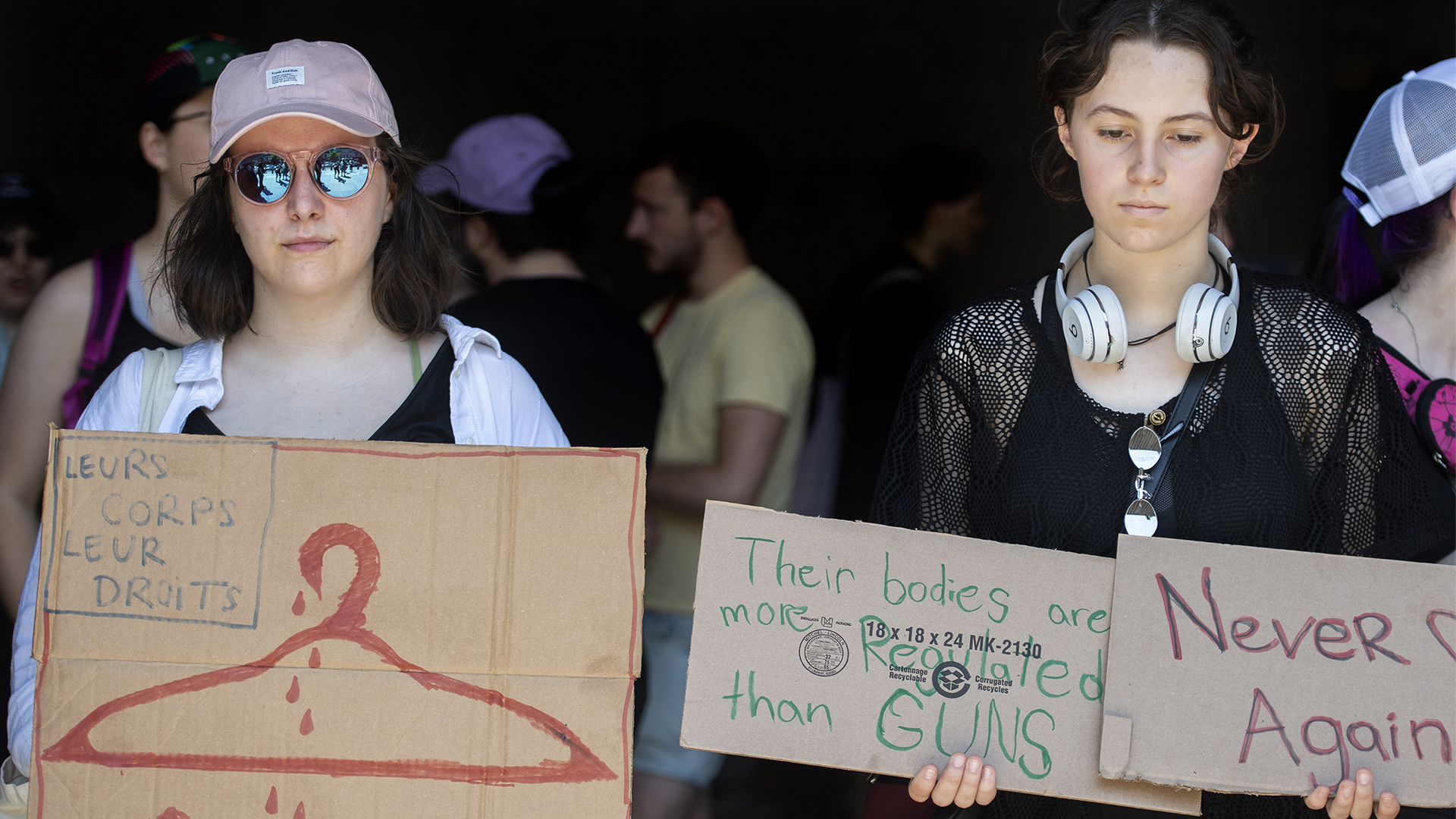 Two young women hold signs that say “Leur corps, leur droits,” “Their bodies are more regulated than guns,” and “Never again.”