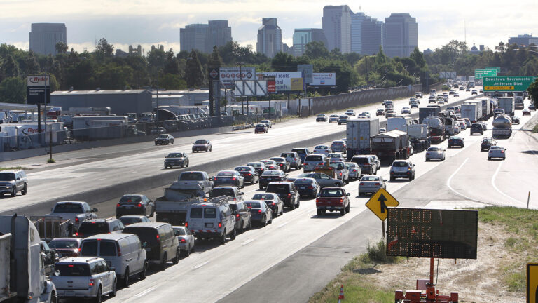 On a four-lane highway, traffic is bumper to bumper in one direction. Bordering the highway are low-rise industrial buildings, a lot with trucks parked, billboards, and in the distance a treed area and high-rise towers beyond them.