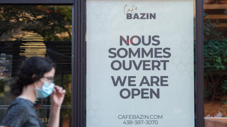 A woman in a grey T-shirt and wearing a mask walks by a sign that says “nous sommes ouvert / we are open” for a coffee shop called Café Bazin.