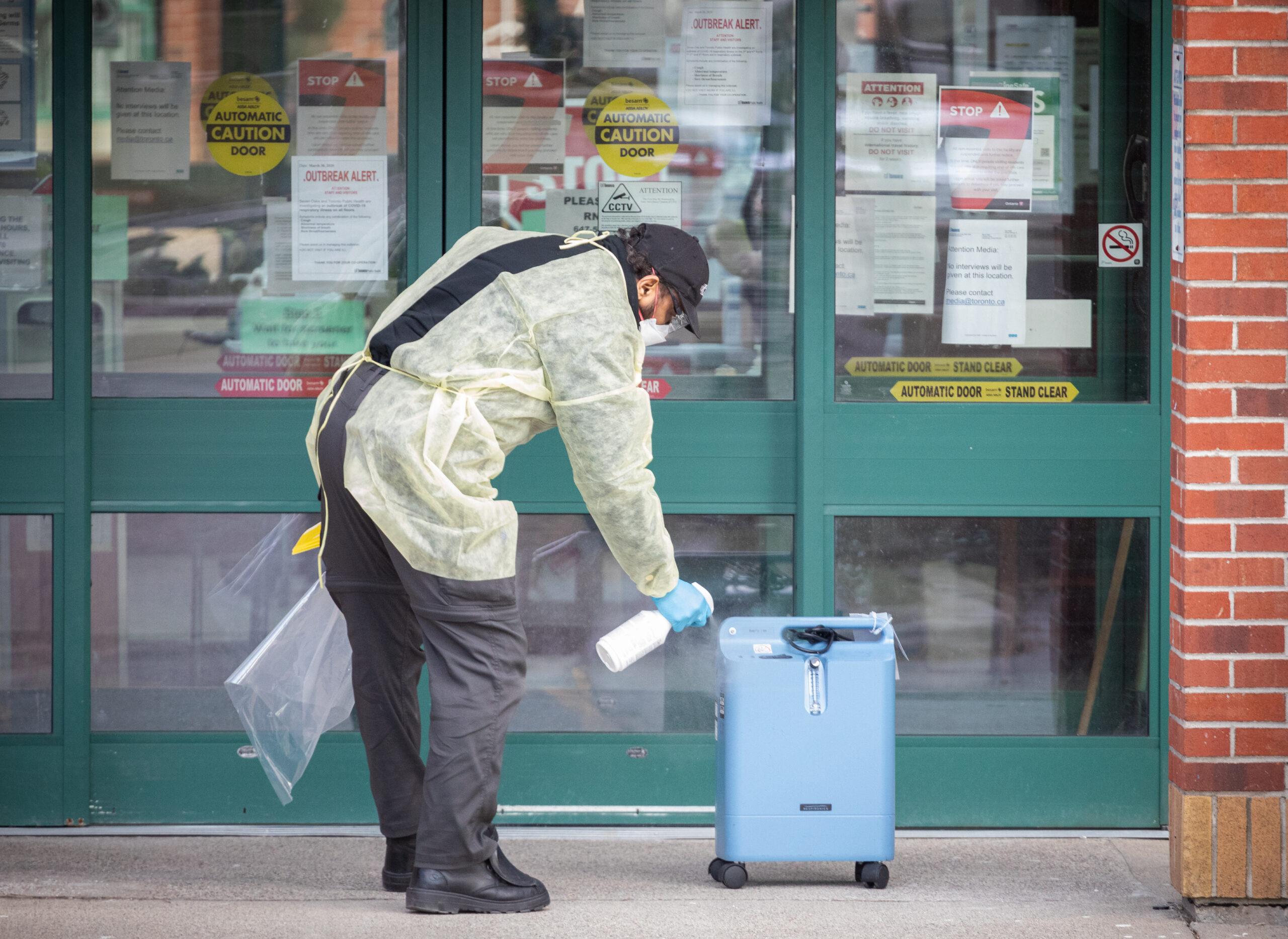 L’homme porte une mince couverture vaporeuse jaune pâle qui se noue autour de ses déchets dans le dos et couvre son front et ses bras. Il utilise un flacon pulvérisateur blanc pour désinfecter un bidon bleu pâle sur roulettes de la taille d’un bagage à main. Il porte une casquette noire, un masque blanc, un pantalon sombre et des chaussures noires, et tient un morceau de plastique dans l’autre main. Il se trouve devant des portes coulissantes peintes en vert. Les vitres sont recouvertes de panneaux d’avertissement.