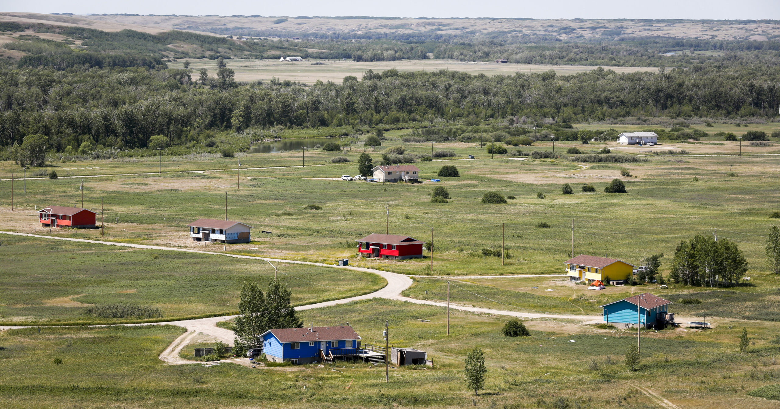A handful of bungalows sit on a cul-de-sac on a flat, grassy expanse of land bordered by a stand of trees, with fields and rolling hills in the distance. The homes are placed far apart from one another.