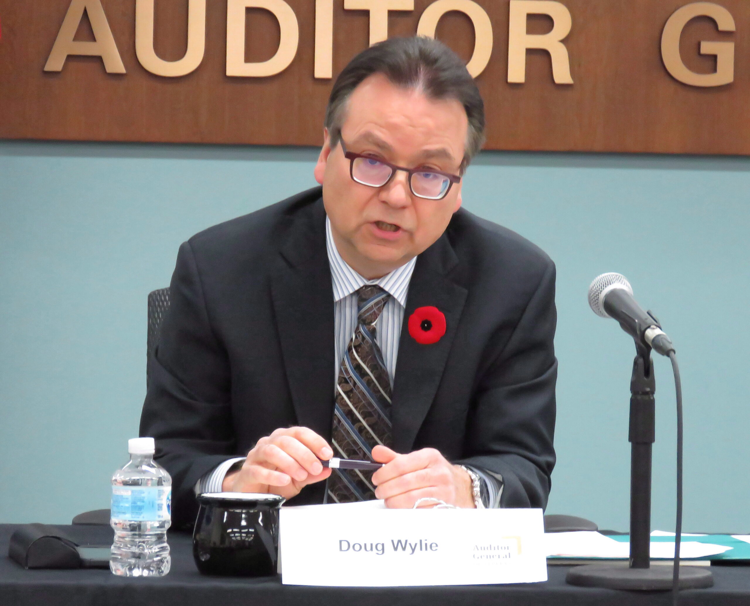 Wylie is seated at a table with a black covering. He is in a dark grey suit with a microphone in front of him. He holds a pen as he speaks. He is middle-aged, white, with short black hair greying at the temples. The wall behind him is teal coloured. High on the wall is a wood sign with the words “auditor general.”