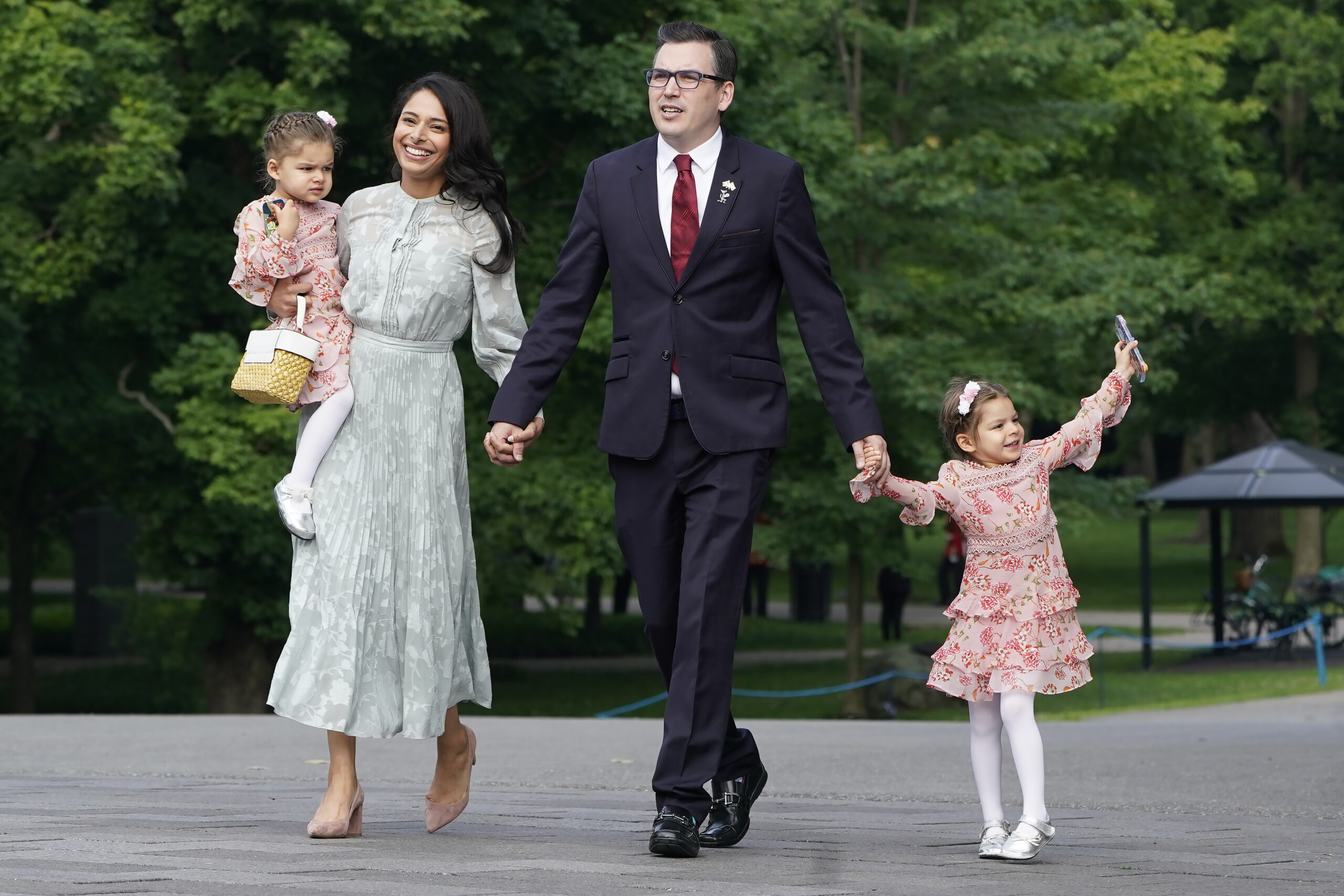 Beech walks along a treed roadway with his wife holding one hand, and one of his daughters holding the other. A second, younger, daughter is carried by Beech’s wife. Beech is in a black suit, white shirt, red tie, black shoes. His wife is in a pale green long-sleeved dress with a high neck and a hem that falls near her calves. Her shoes, a low heel, are nearly invisible they match her skin colour so closely. The two daughters are in pale pink dresses that fall at the knee, with white tights and white shoes.