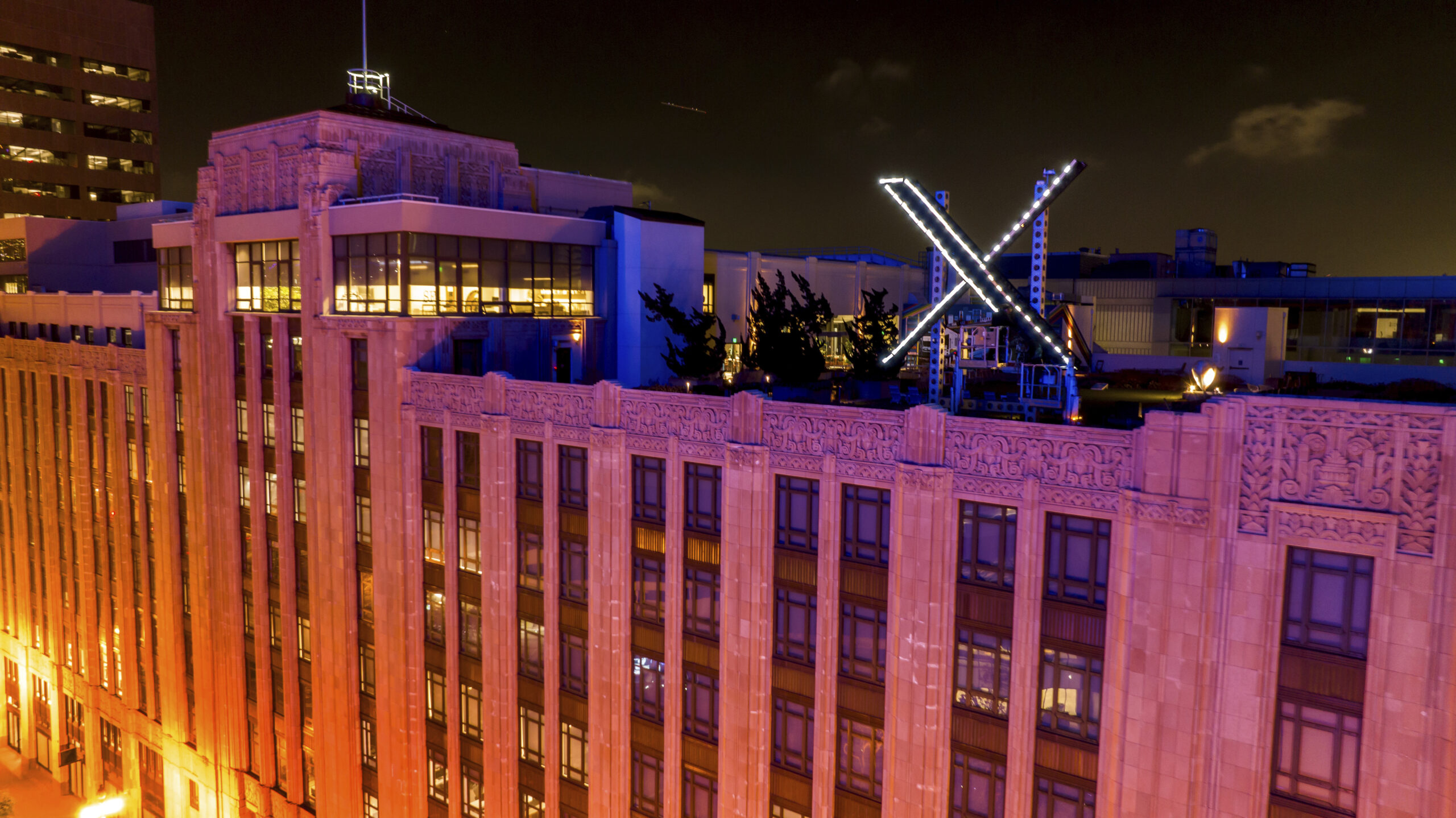 The building is six floors high, and ornate in style, made of smooth stone, decorative carvings along the top, with black-framed windows. It’s night-time, and a bright light bathes the building in tones of yellow, orange and magenta. The rooftop has evergreen trees and a large lit-up “X” that is black with white lights.