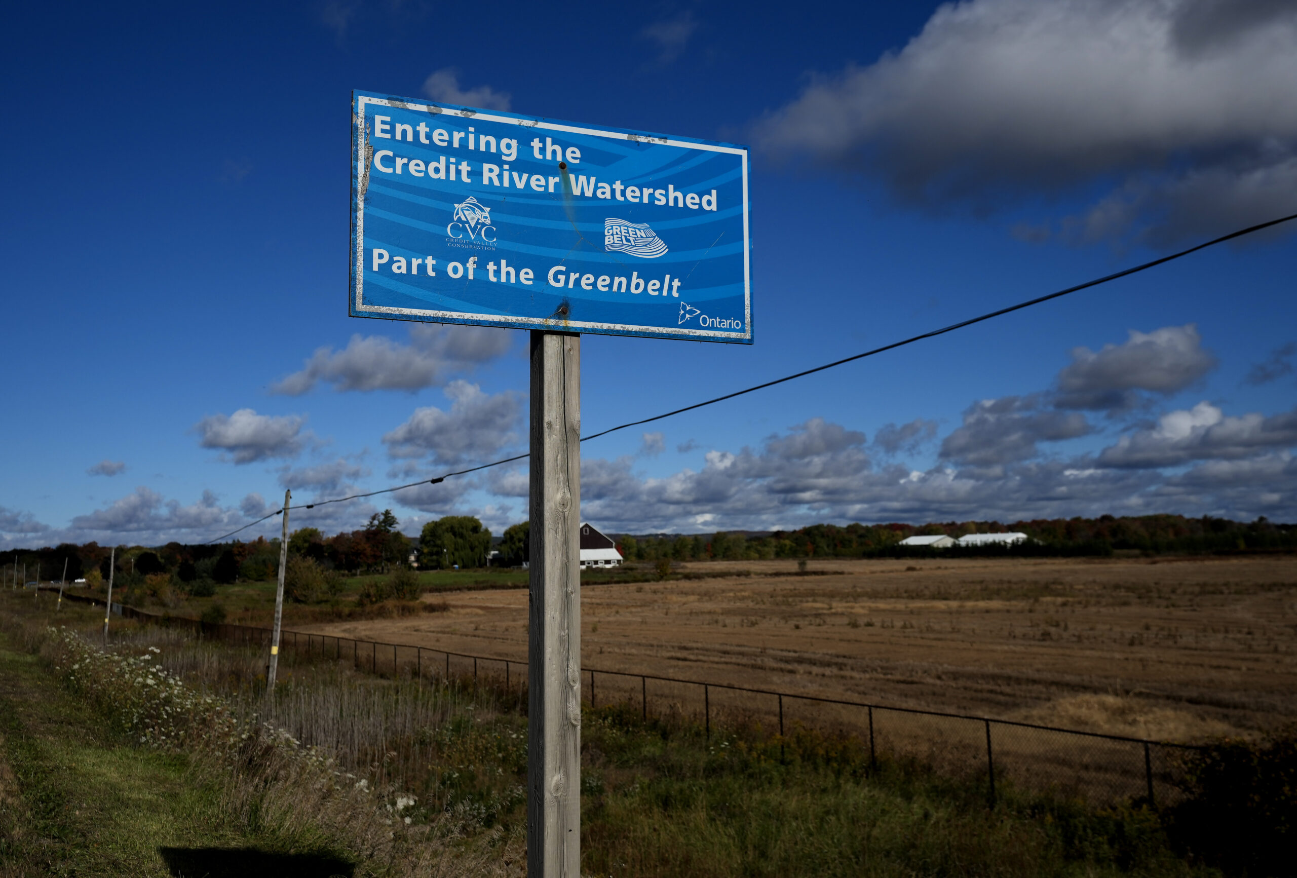The sign is blue and says “Entering the Credit River Watershed. Part of the Greenbelt”. The road is lined with tiny wildflowers that border a swath of tall grass and a fenced-in field that has turned golden yellow. In the distance are a white farmhouse and a few white one-storey buildings on the property. The sky is a deep blue with low cloud cover near the horizon.