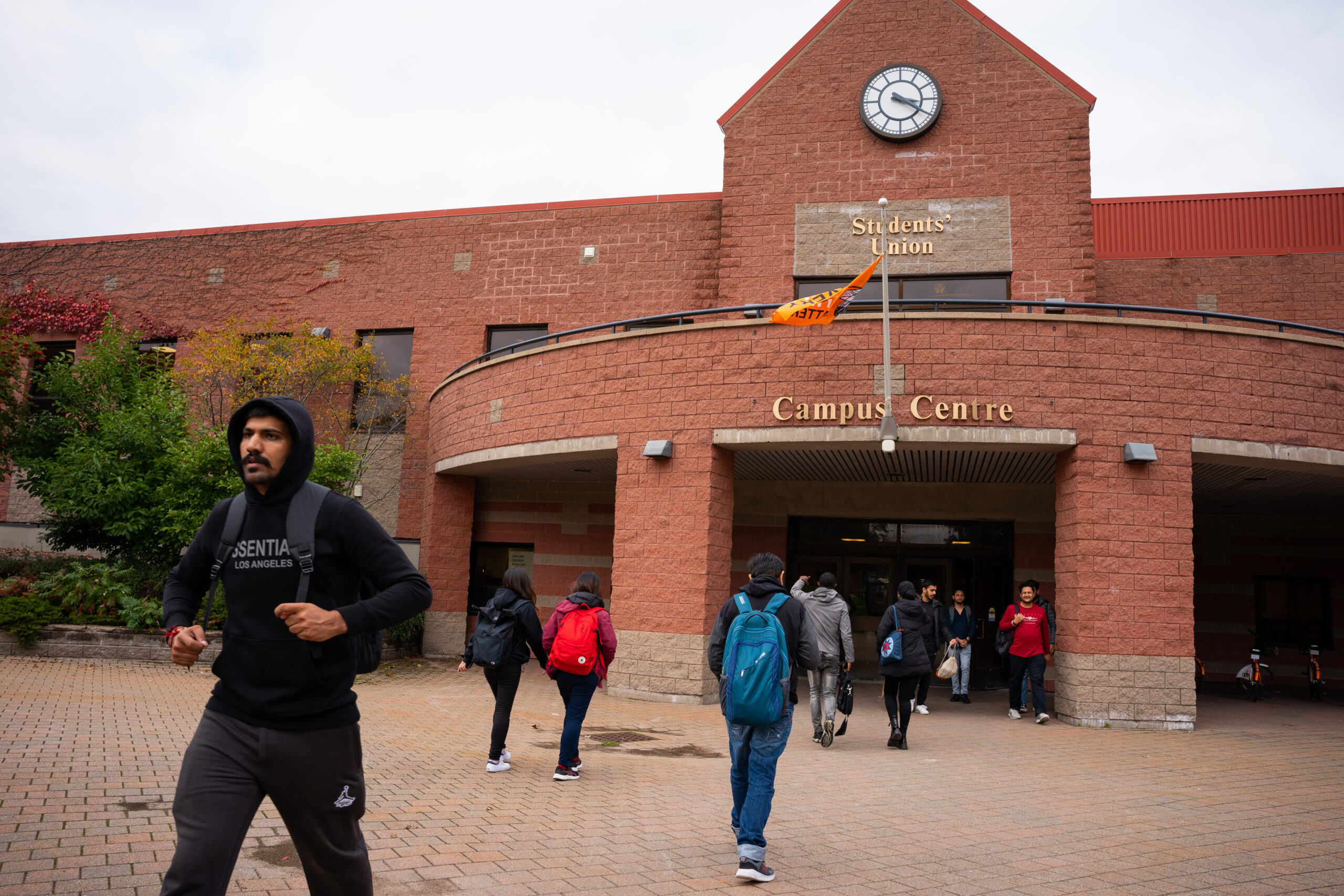 About eight students exit and enter a wide entrance to a two-storey brick building surrounded by a courtyard with a few trees.