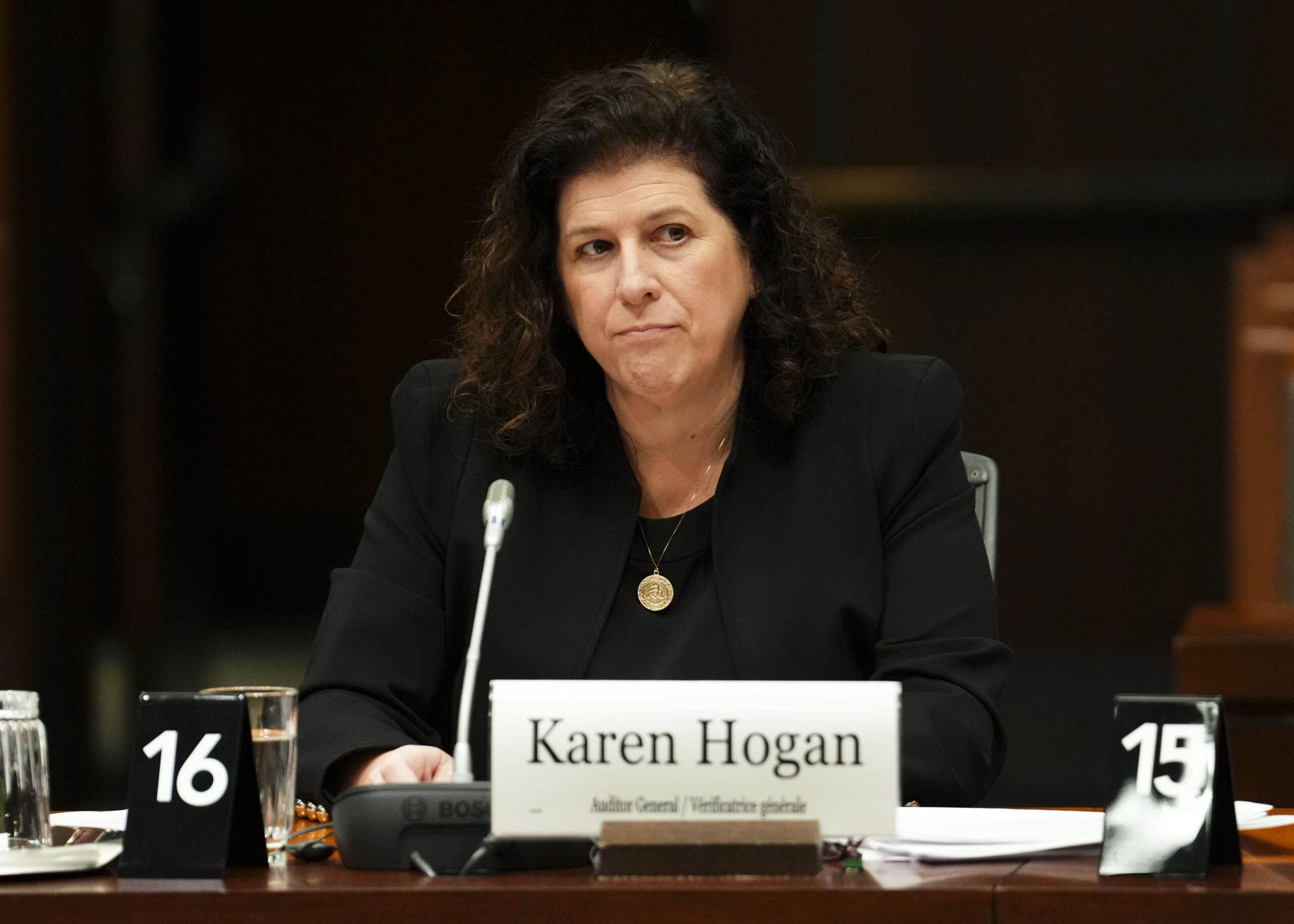 Mme Hogan est assise à une longue table en bois, avec un microphone et un badge devant elle. Elle porte un blazer et une chemise noirs et écoute d’un air sérieux.