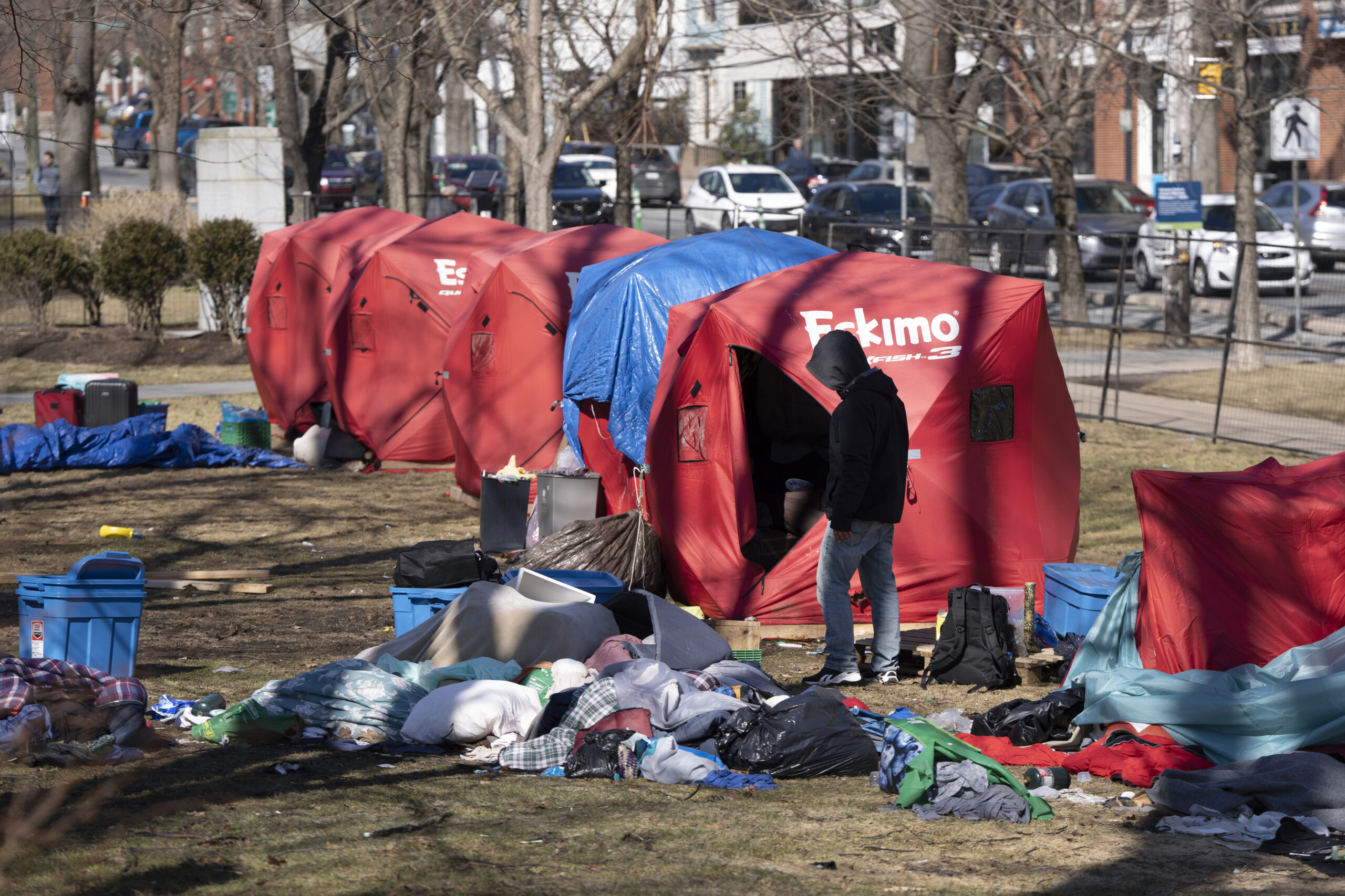 Four red tents sit side by side, with a blue tarp tied over the top of one of them. Piles of clothing and plastic bins are strewn on the ground.