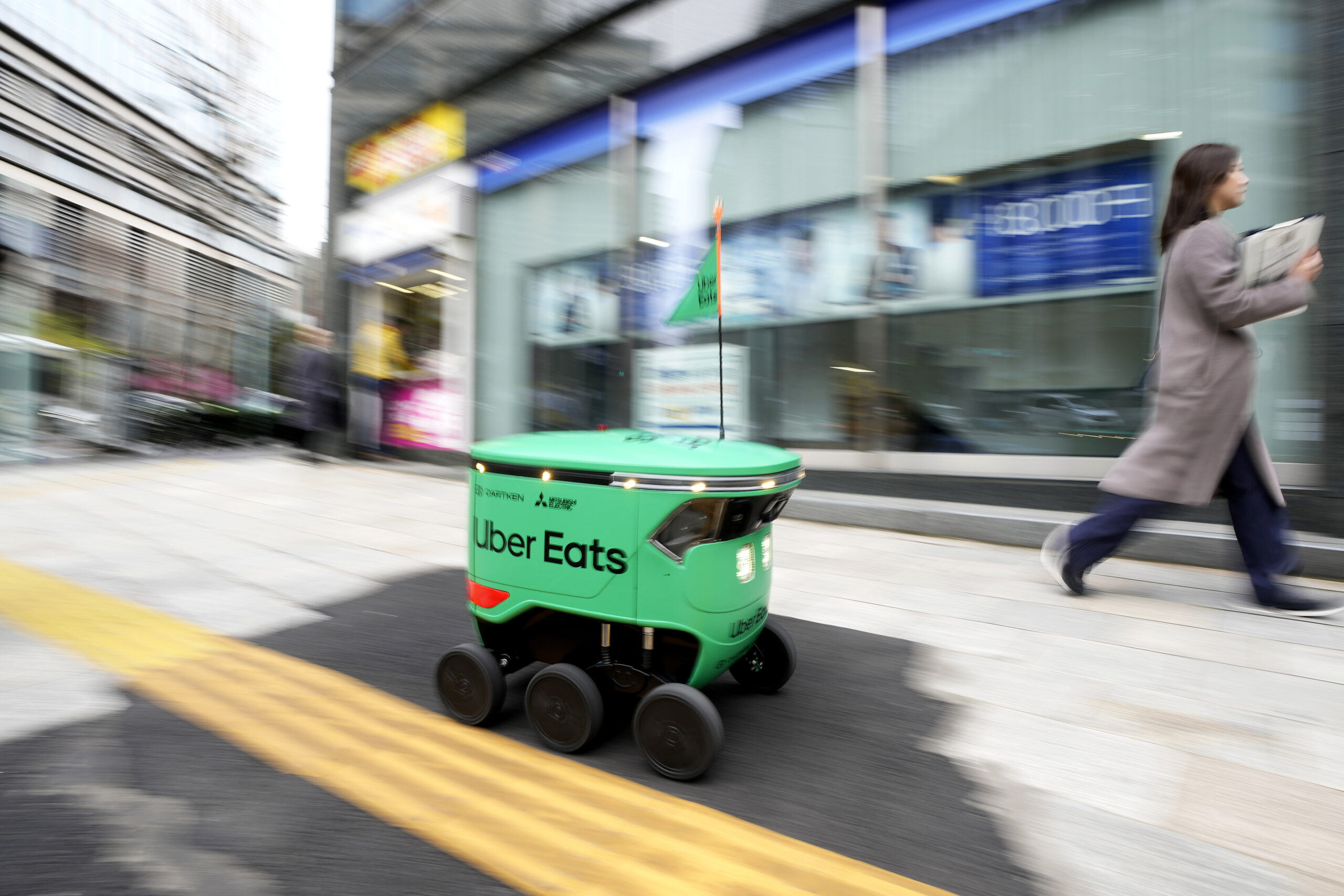 The robot is a compact, mint-green enclosed container with rounded edges, three black wheels on both sides, and a flag pole bearing a small triangle of green. It is moving a few feet away from a woman walking in the same direction. The photographer has panned so that the robot is in focus while the sidewalk and the nearby buildings are blurred.