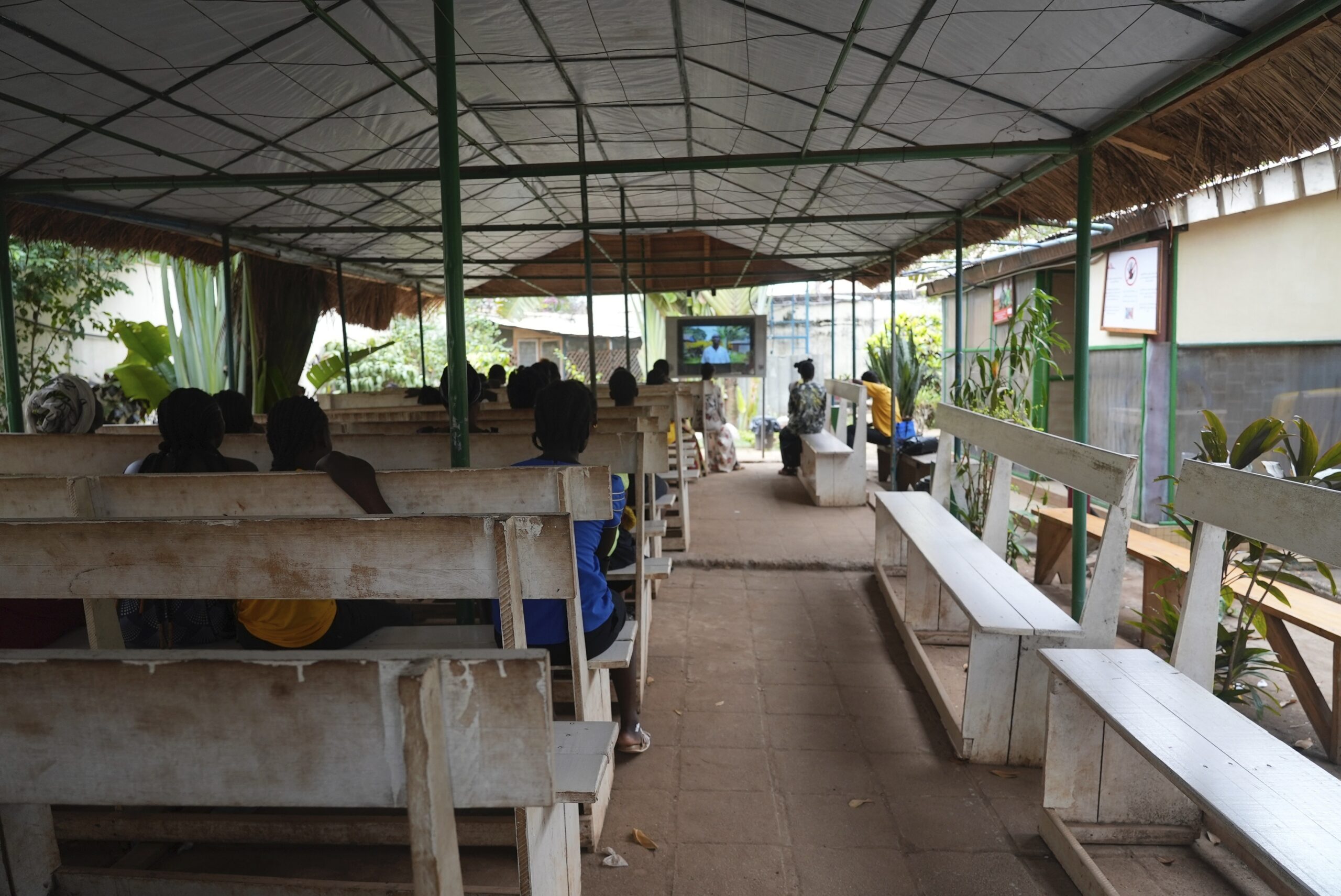 In an open-air metal structure with a thatched roof, women sit in rows of wooden benches. They are all looking at a computer or TV screen at the front of the room.