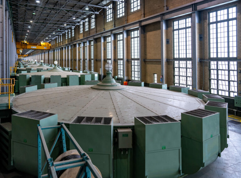 The building has a long corridor with high ceilings, brick walls, two storeys of tall windows with many mullions, and a ceiling with metal pieces criss-crossing. Nine of the metal rotors fill the space. Each one is large and round, with green metal boxes lining the edge. They look like large spinning tops.  