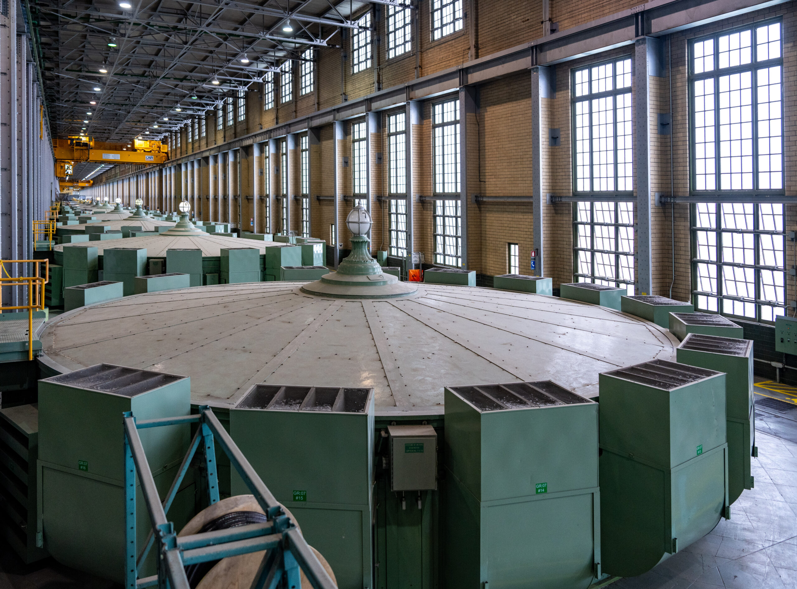 The building has a long corridor with high ceilings, brick walls, two storeys of tall windows with many mullions, and a ceiling with metal pieces criss-crossing. Nine of the metal rotors fill the space. Each one is large and round, with green metal boxes lining the edge. They look like large spinning tops.