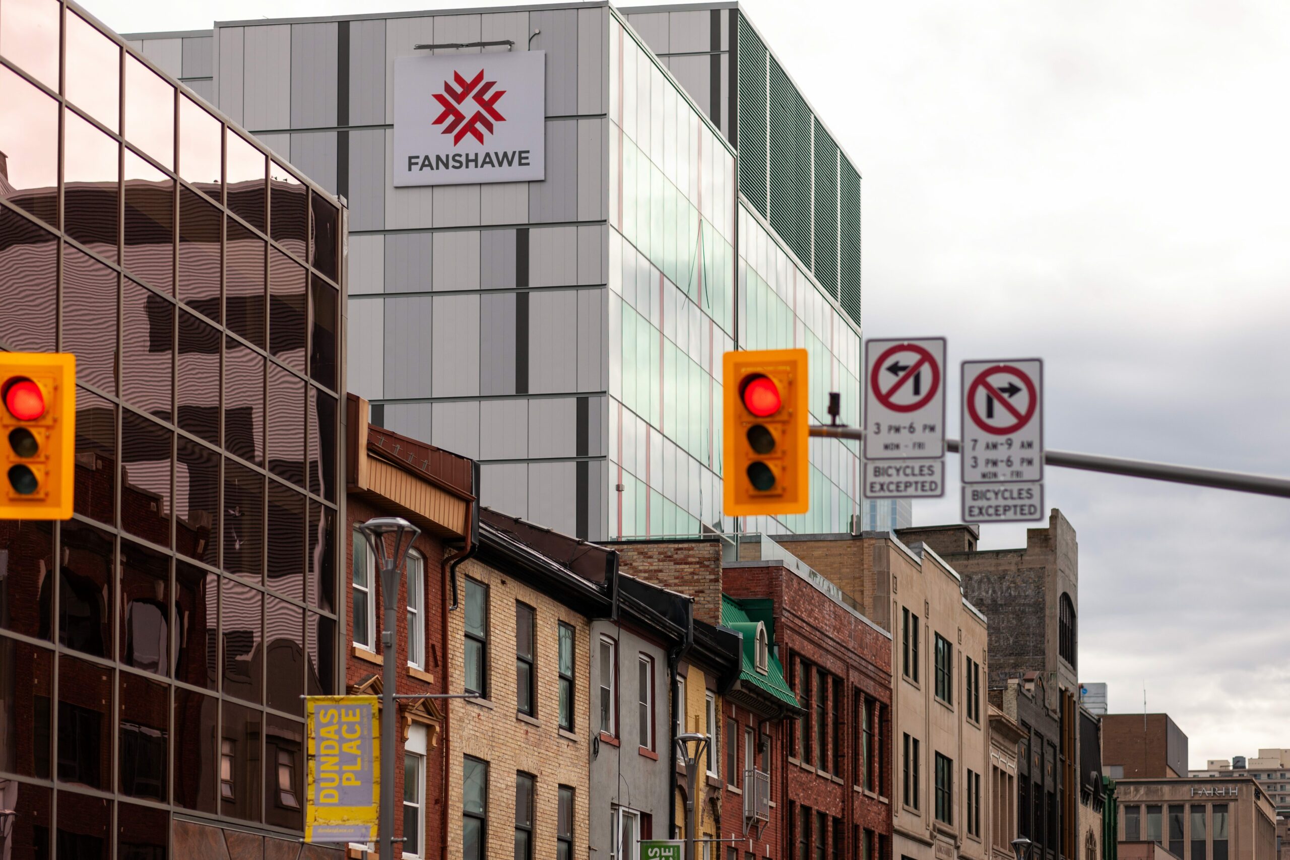 On a street of older brick buildings several storeys high, a glass addition has been built at the rear of one property. It is several storeys higher and has a square white sign that says Fanshawe with a red logo of chevrons in a circle.