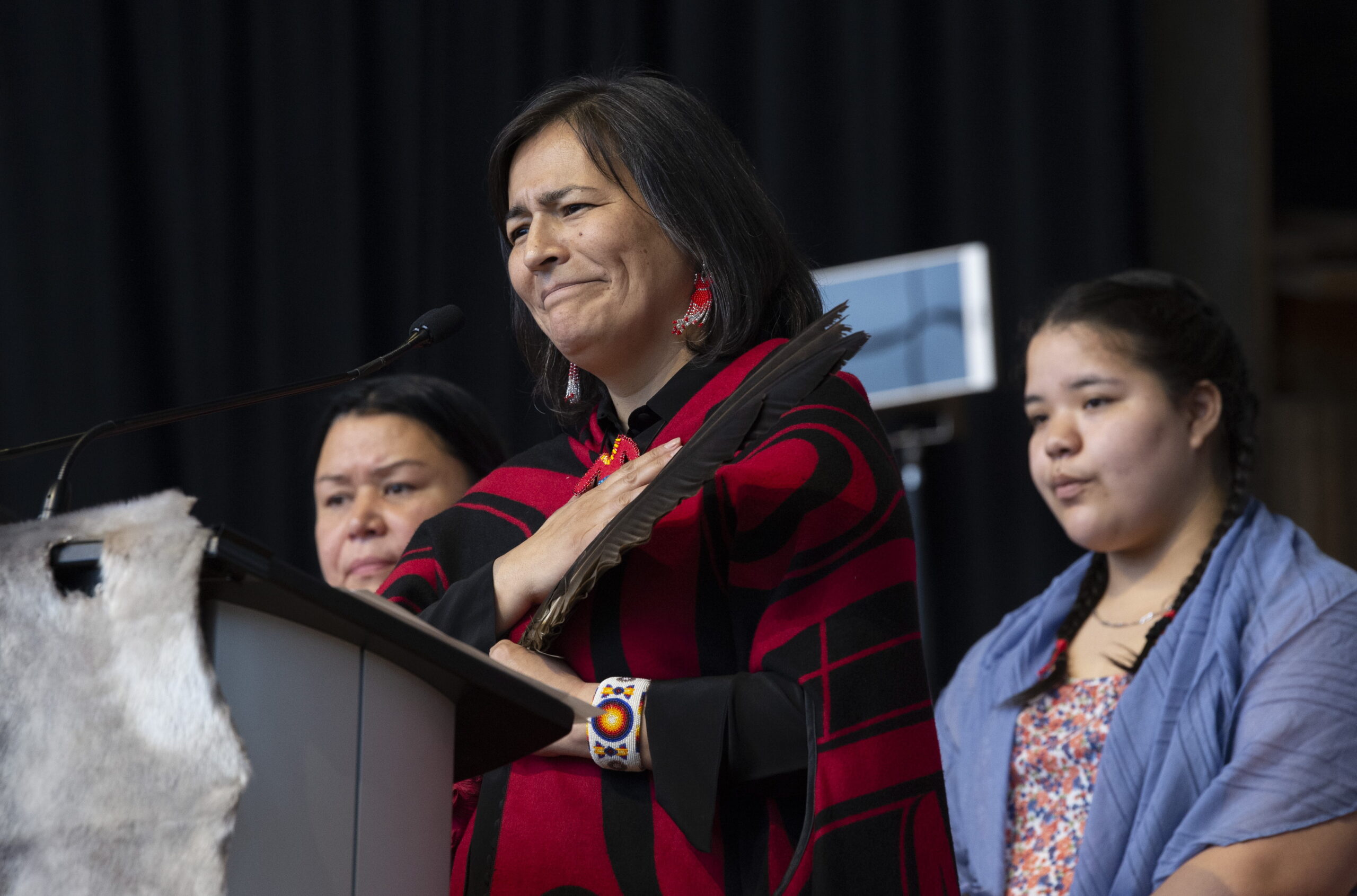 Audette stands at a podium dressed in red and black with a hand placed over her heart and in her other hand a dark feather. A middle-aged woman and a young woman stand on either side of her. Draped on the front of the podium is a light-grey animal hide.