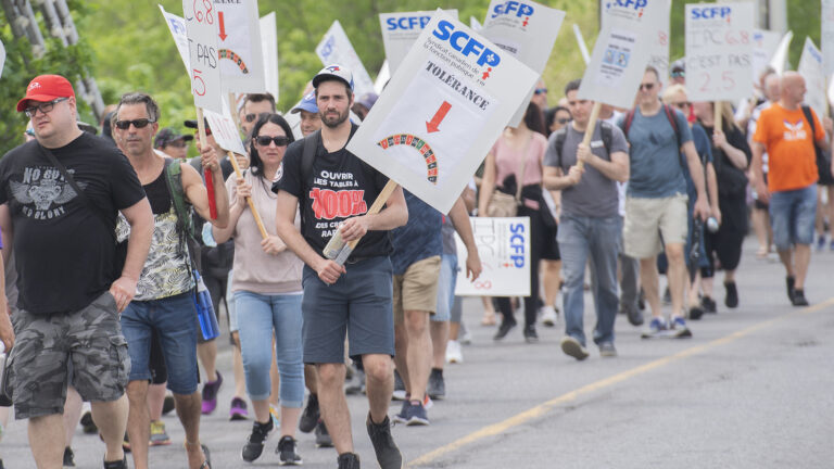 A large group of men and women in t-shirts, shorts and jeans walk down the road. Some of them carry signs.