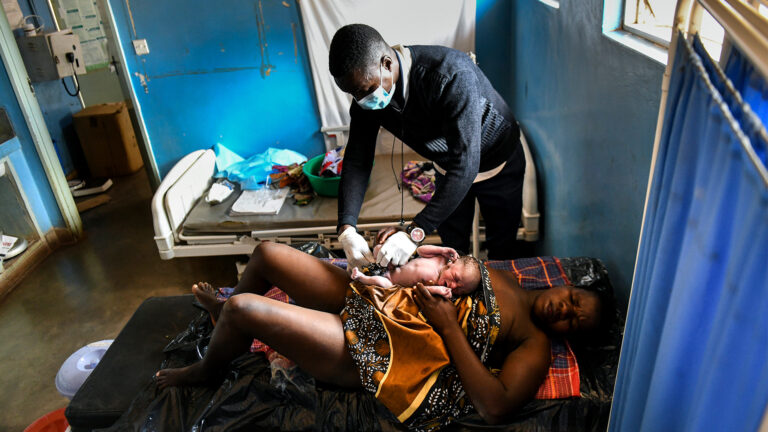 In a small room with blue walls, two simple hospital beds, two small windows, and a white sheet tacked to the wall, a woman lies with her eyes closes, head turned sideways, on a bed with a black mattress, her new baby resting on her tummy. She looks exhausted. A patterned cloth with browns, whites and black covers her midriff, with the baby on top of it. Her knees are up and her legs are bare. The midwife, a man, wears white rubber gloves as leans over to clean the baby.