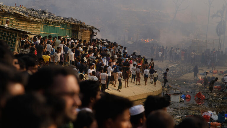 Smoke fills the air as hundreds of people stand clustered on an incline next to a series of shacks with fabric for walls. In the distance, the smoke is thicker, with small fires burning among palm trees seen in silhouette.