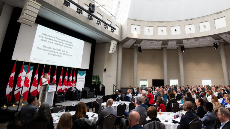 In a cavernous rood with a domed ceiled and stone pillars lining the walls, Joly speaks at a lectern as she addresses a seated crowd of people at round tables. Behind Joly is a row of 11 Canadian flags draped on poles. Above her is a large screen with written info.