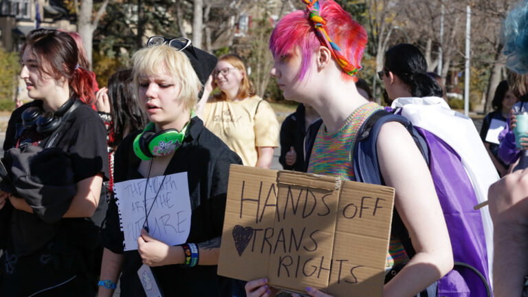 A youth with pink hair holds a sign saying “Hands off trans rights” next to another youth with blonde hair with a sign saying “the youth are our future.” Other protesters are in the background.