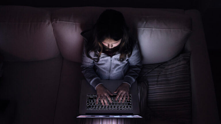 An unidentifiable child types on a laptop computer while sitting on a couch.