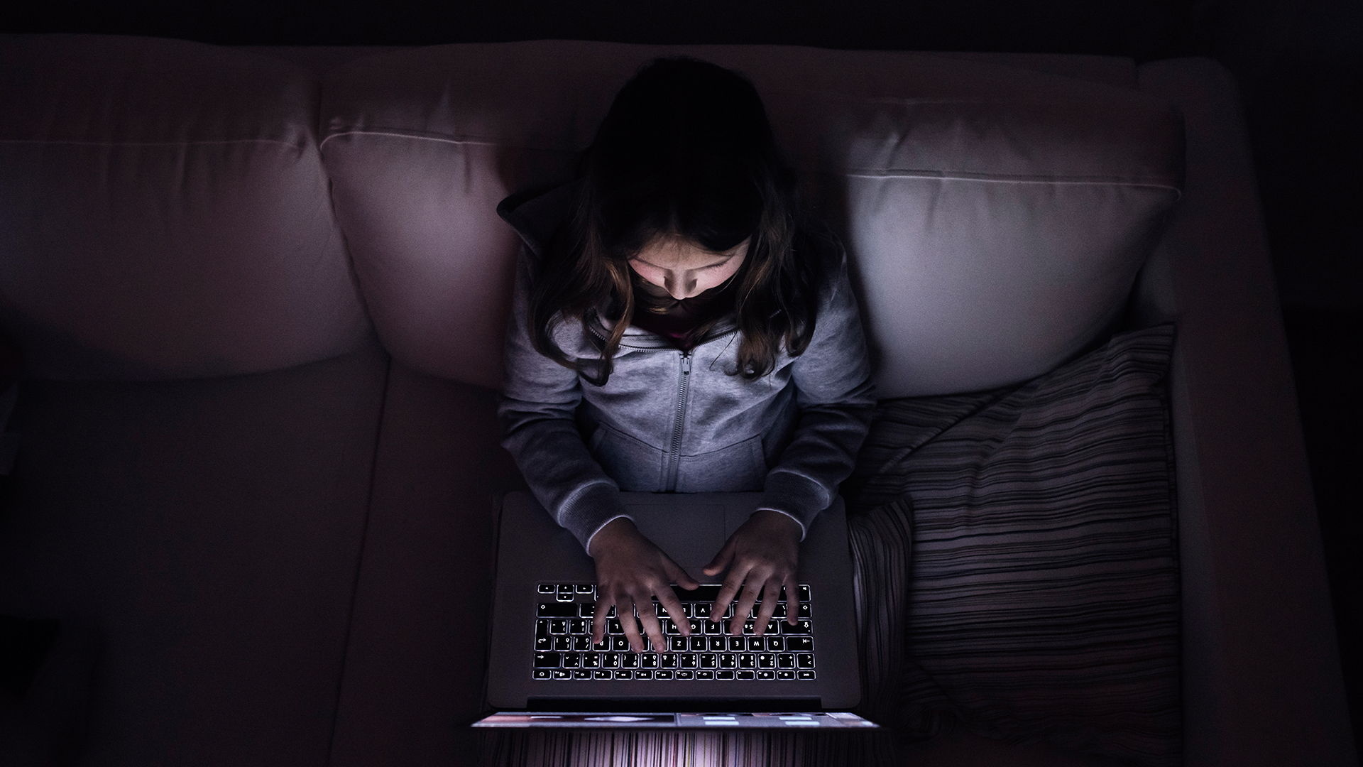 An unidentifiable child types on a laptop computer while sitting on a couch.