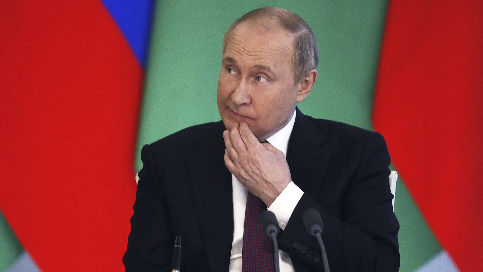 Vladimir Putin scratches his chin and looks to the side while he stands at a podium with the flags of both countries hanging behind him.