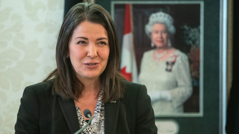 Danielle Smith parle au micro alors que l'on voit un portrait de la reine Elizabeth II en arrière-plan.
