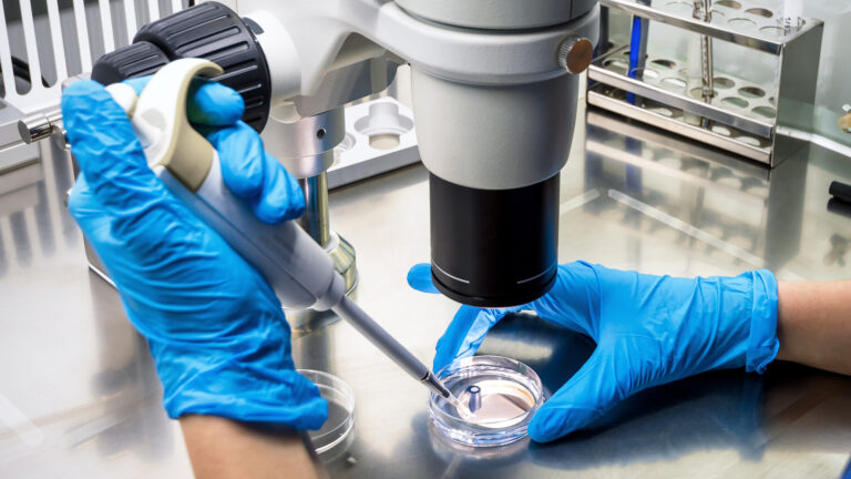 In a lab, a pair of hands in blue latex gloves holds an instrument that drops liquid into a clear shallow, round piece of plastic. Above the specimen is a machine with a tube aiming down. A rack holding several small tubes sits nearby.