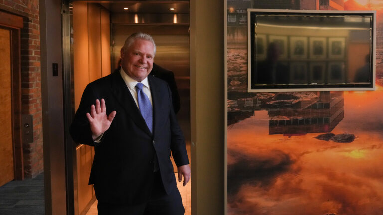 Doug Ford waves at a photographer as he walks into a room with brick walls and a monitor on the wall.