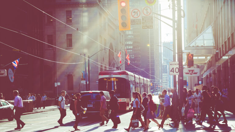 A dozen men and women cross at a downtown intersection in Toronto’s downtown, surrounded by tall buildings as cars and a streetcar drive past on a sunny day.