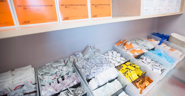 Two long shelves filled with bins of small packets and plastic containers along with boxes of syringes.