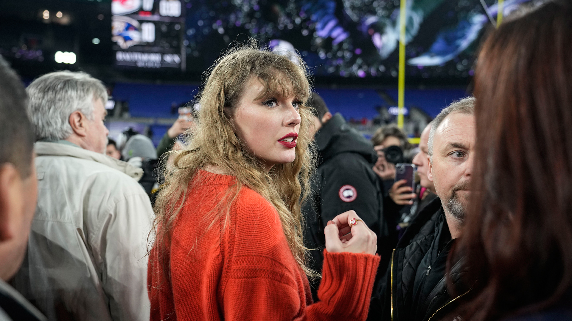 A photo of Taylor Swift in a red sweater, appearing to look for someone, surrounded by a crowd of people in a football stadium.