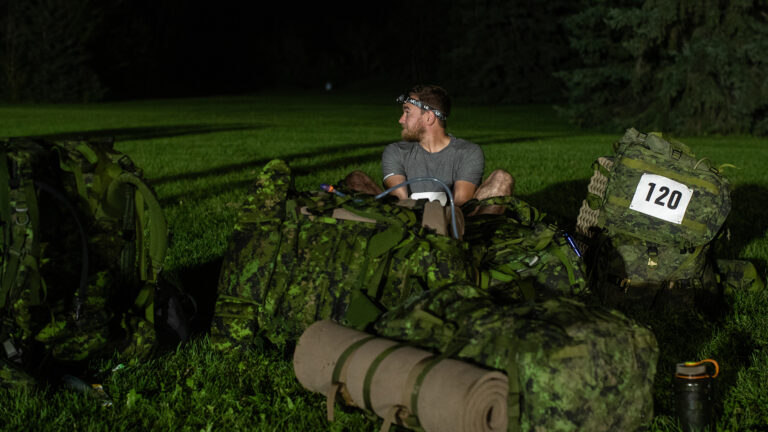 Captain Chris Knauer sits in a large lawn under a dark sky wearing a headlamp among camouflage green backpacks.