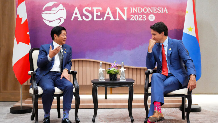 The two men sit opposite each other, separated by a small dark wood accent table with a pot of flowers and two water bottles. Behind them are two flags and a purple and pink backdrop that says ASEAN, Indonesia 2023.