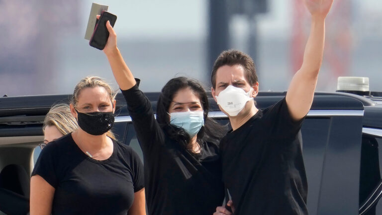 Kovrig and his wife both have one arm raised as they acknowledge their arrival, standing with his sister in front of a black SUV on the tarmac where the jet carrying him to Canada arrived. All three are wearing masks.