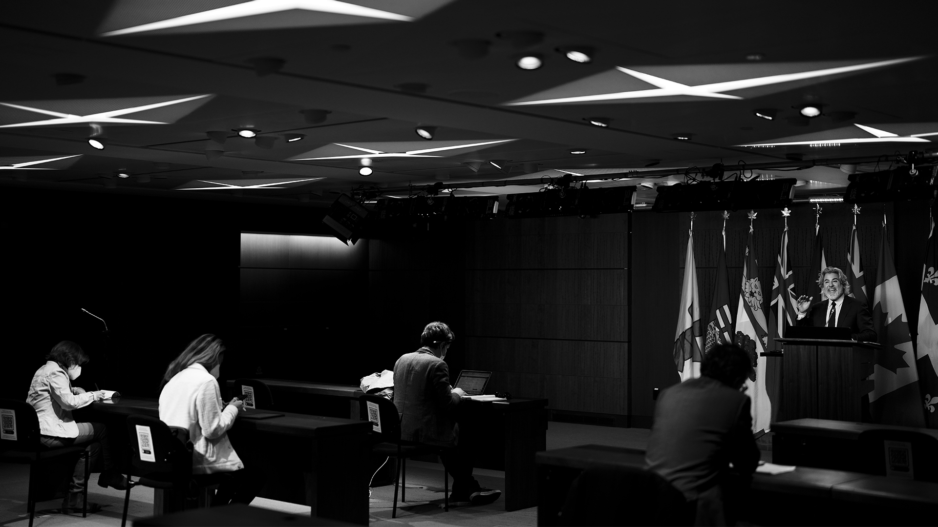 A black-and-white image of reporters at tables listening to Heritage Minister Pablo Rodriguez.