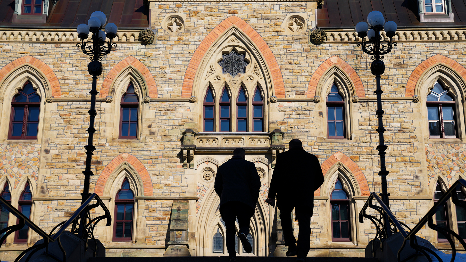 Two men in silhouette walk up the steps leading to a large, three-storey building with ornate architectural details including arch-shaped windows and brickwork detail around them.