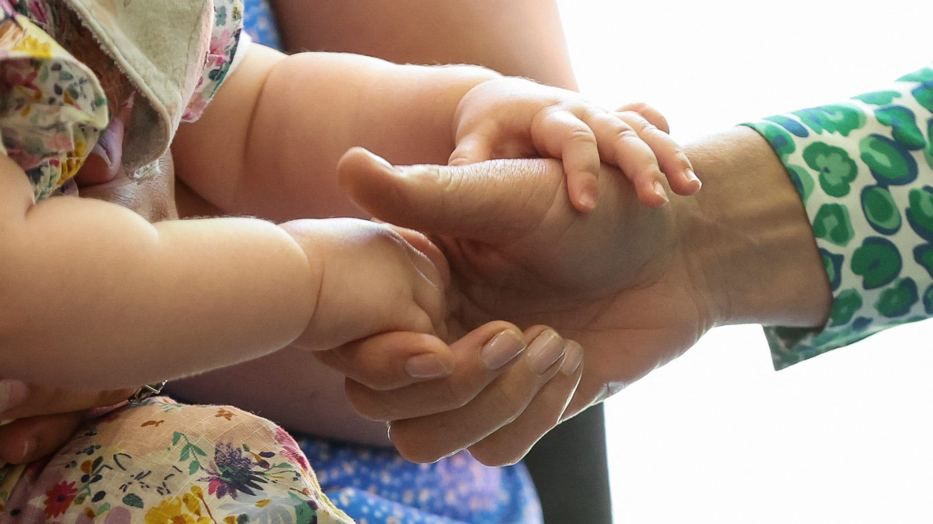 A closeup of an adult holding a baby while a hand reaches in to hold the baby’s hand.