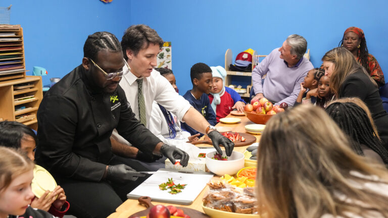 The prime minister has the sleeves of his white dress shirt rolled up. He’s wearing black gloves while reaching for strawberries from a bowl. Several children seem bemused.