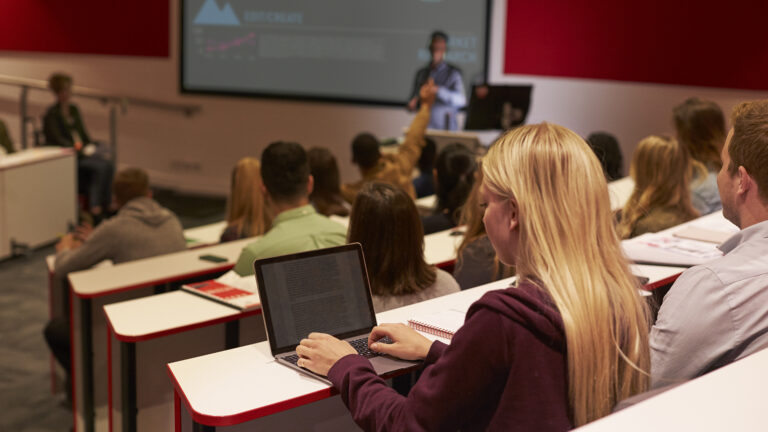 Students sit in a classroom with a teacher standing in front of a large screen. The rows of seating are tiered. Some of the students are using laptops.