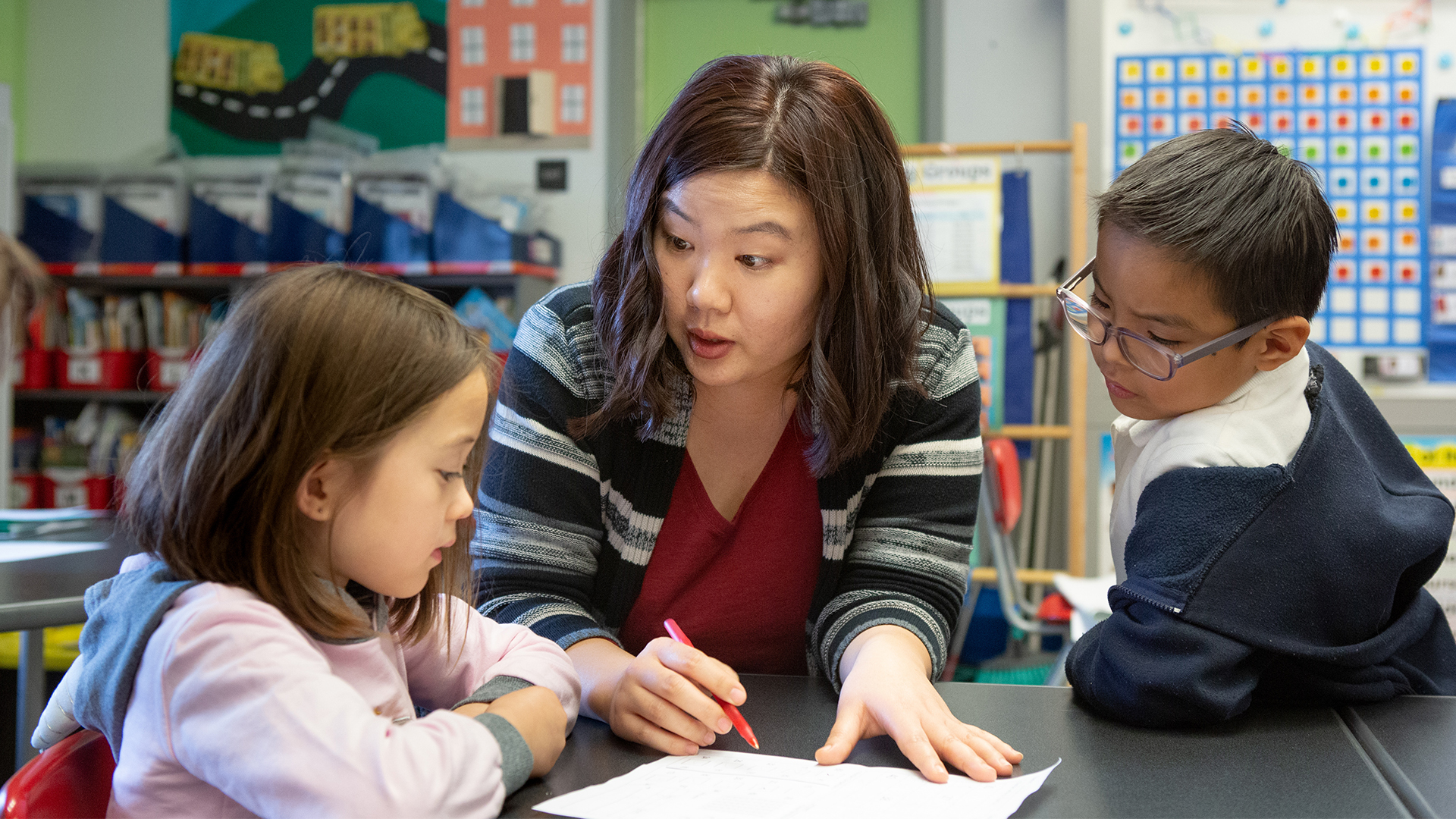 A kindergarten teacher helps a boy and girl with a class activity. They are looking at a piece of paper on a table in front of them while she is speaking with a red pen in hand.
