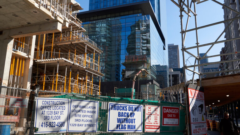 Next to a tall glass tower, the exposed storeys of a new tower are going up. The concrete floors are held up by temporary orange-coloured steel supports. The metal fence around the project has five signs identifying the construction company and warning trucks not to back up without a flag man. Directions for an on-site defibrillator are there as well as warning signs.