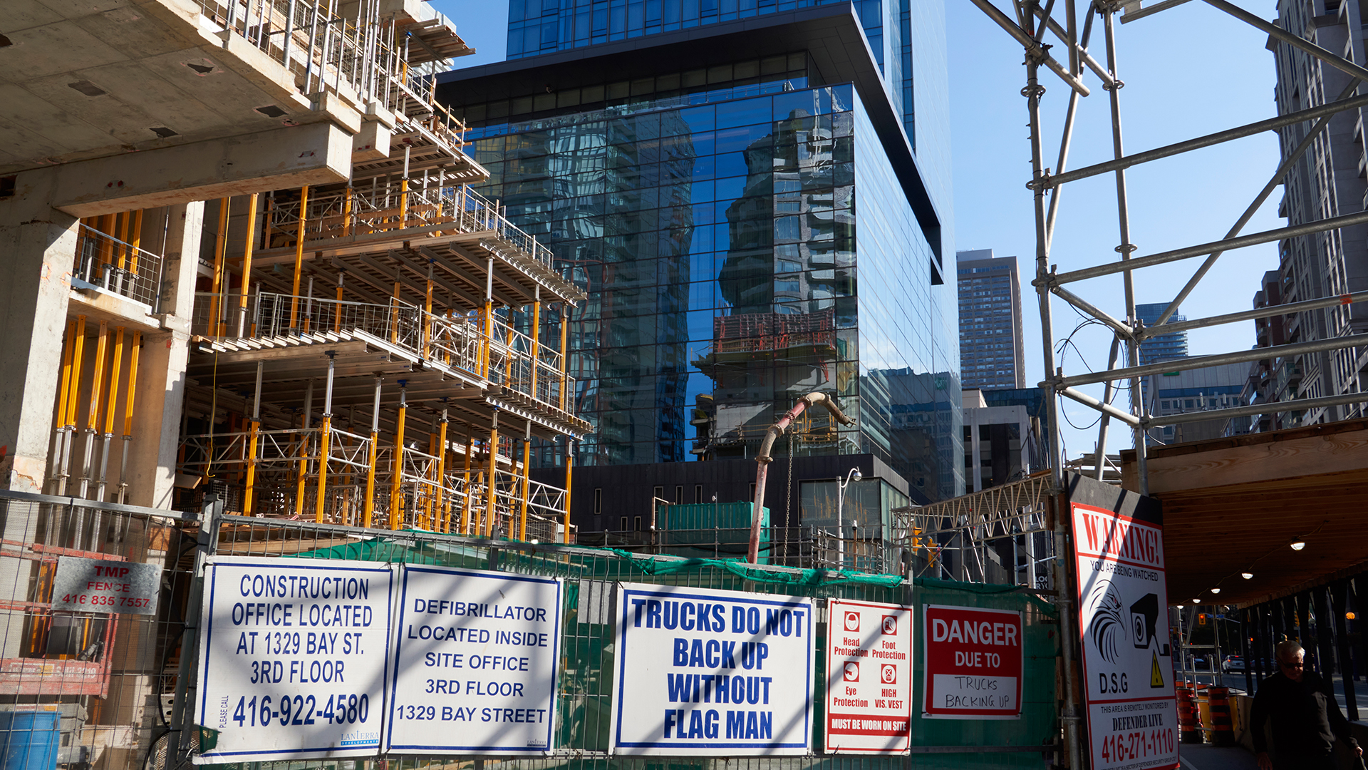 Next to a tall glass tower, the exposed storeys of a new tower are going up. The concrete floors are held up by temporary orange-coloured steel supports. The metal fence around the project has five signs identifying the construction company and warning trucks not to back up without a flag man. Directions for an on-site defibrillator are there as well as warning signs.