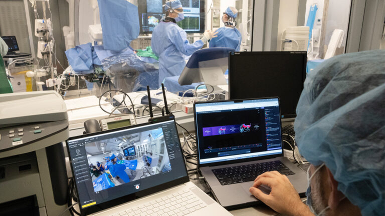 Two laptops and other electronics are set up in a room adjacent to the operating room. A wide window pane separates the two spaces. At the laptops sits a man in blue scrubs, his hand on one of the laptops. On the other side of the glass, two male doctors talk as they look at an oversized monitor on the wall that shows graphs and charts. Next to the doctors is a patient on a gurney.